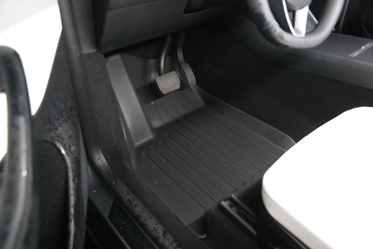 Close-up of the driver-side footwell in a 2026 Tesla Model Y (Juniper) featuring LinerX all-weather black floor mats, accelerator and brake pedals, and part of a white seat.