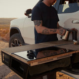 A person with tattoos pours hot water from a kettle on the Thunderbolt Adventure Supply Rivian R1T Overland Kitchen with updated Gear Sled, set up on a white vehicle outdoors at sunset.