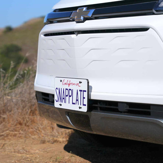 Close-up of a Chevrolet vehicles front end with a California license plate reading SNAPPLATE in purple on white. The vehicle is parked outdoors.