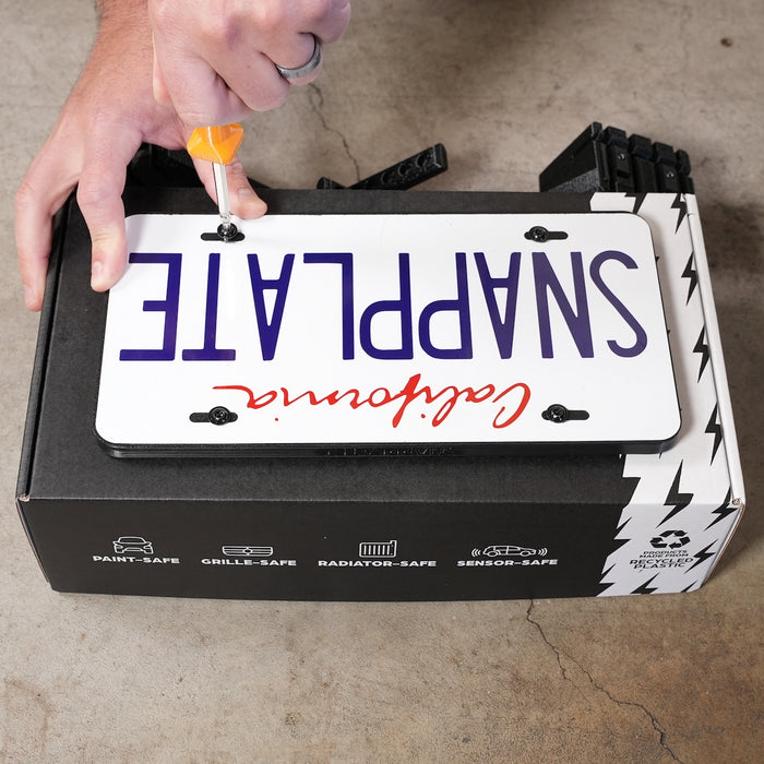 A person uses a screwdriver to attach a novelty license plate reading SNAPPLATE upside down on top of a black box with automotive icons and text.