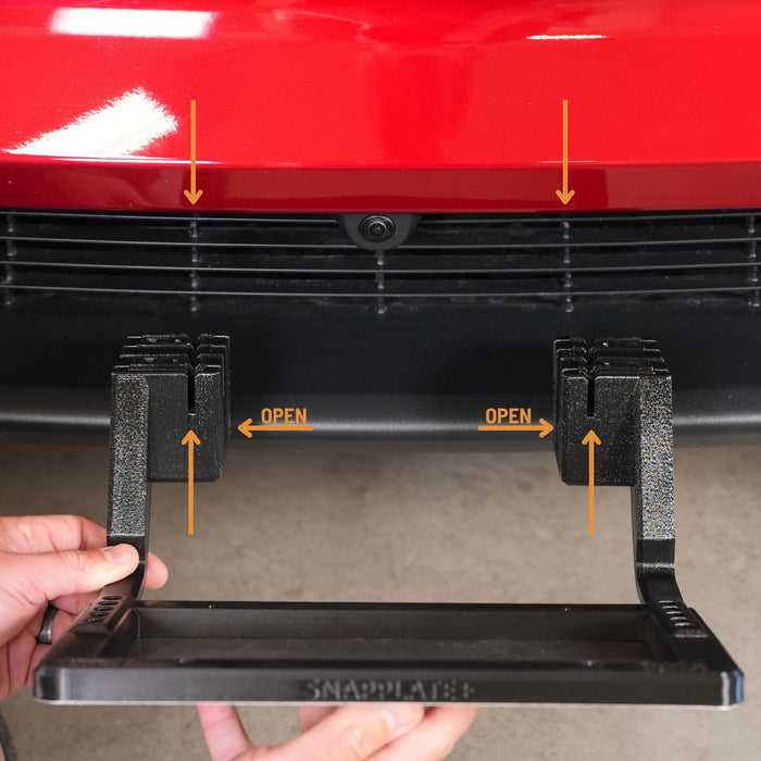 A person holds a license plate mount near the front of a red vehicle, with arrows indicating alignment points and labeled open on the mounts hinges.