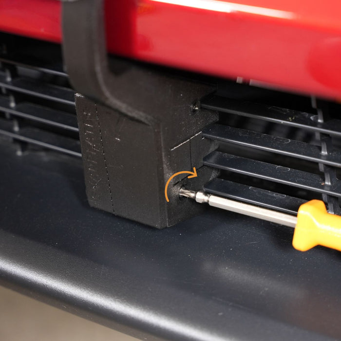 A screwdriver is turning a screw to attach a black plastic piece onto the front grille of a red vehicle.
