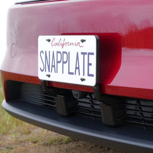 Close-up of a California license plate reading SNAPPLATE mounted with the EveryAmp Tesla Model Y SnapPlate Front License Plate Holder on the grille and bumper of a red Tesla Model Y.