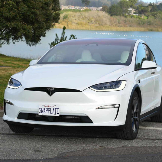 White Tesla Model X parked by a lakeside with trees in the background; the license plate reads SNAPPLATE.