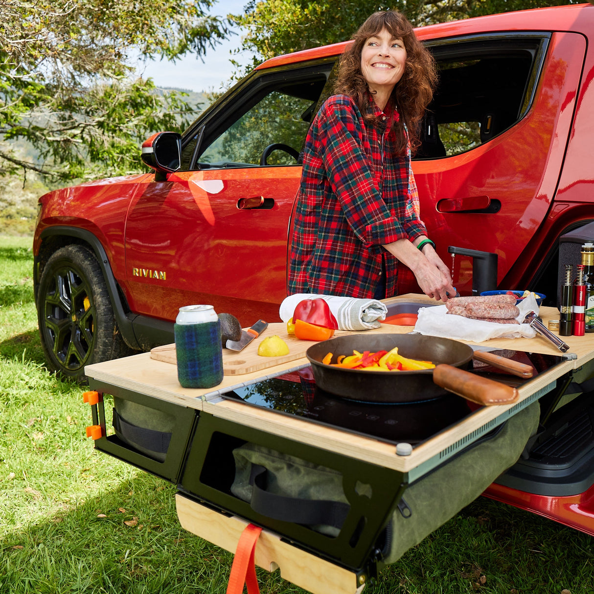 A person cooks outdoors on a Thunderbolt Adventure Supply Rivian R1T - Overland Kitchen with updated Gear Sled, attached to a red SUV, using ingredients and a skillet, creating an ideal campsite cooking setup on the grass.