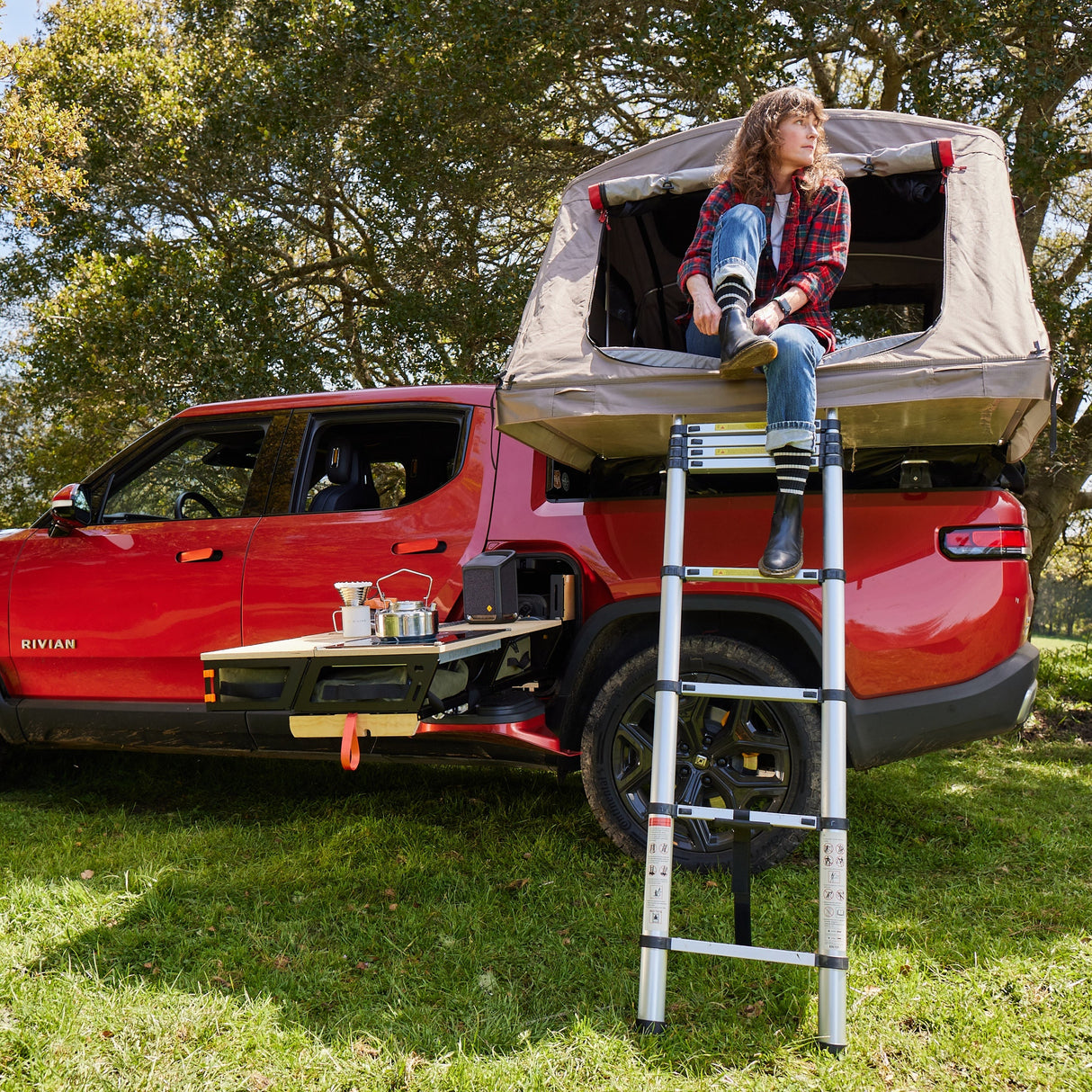 A person sits at the entrance of a rooftop tent on a red Rivian R1T equipped with Thunderbolt Adventure Supply's Overland Kitchen and updated Gear Sled, parked on grass under trees and ready for adventure.