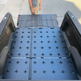 A person stands near the open tailgate of a pickup truck equipped with the Thunderbolt Adventure Supply R1T Bed Glider, showcasing its black, perforated bed cover and organized storage system in the bed.