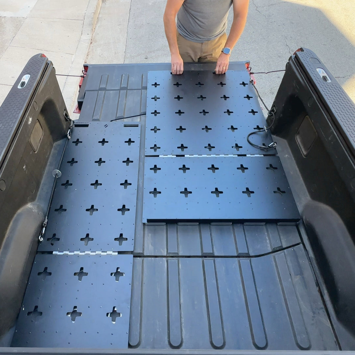 A person installs modular black panels with cross-shaped holes in the Thunderbolt Adventure Supply R1T Bed Glider, optimizing pickup truck bed storage for camping accessories.