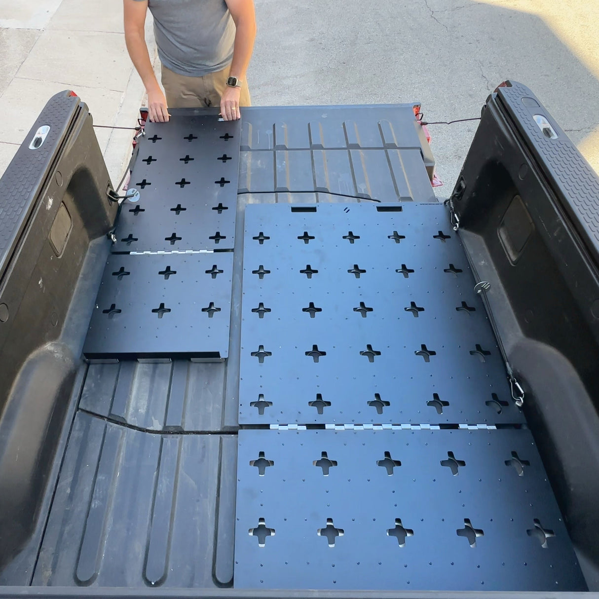 A person installs Thunderbolt Adventure Supply’s R1T Bed Glider—modular black panels with cross-shaped holes—into a pickup truck bed, creating versatile storage for camping accessories.