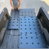A person installs Thunderbolt Adventure Supply’s R1T Bed Glider—modular black panels with cross-shaped holes—into a pickup truck bed, creating versatile storage for camping accessories.
