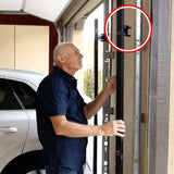 An older man inspects a garage door track in a garage; a red circle highlights The Infinity Shield Infinity Shield Brackets and the garage door sensor mounted on the vertical rail near the top.